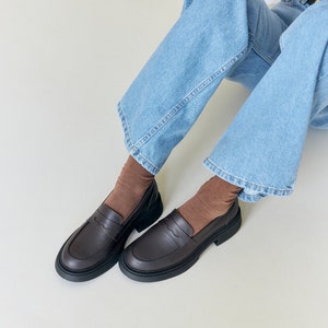 K&ouml;nnte beinhalten: Dunkelbraune Leder-Loafer mit dicker schwarzer Sohle. Die Schuhe haben ein klassisches Penny-Loafer-Design. Die Schuhe werden mit braunen Socken und hellblauen Jeans getragen. Die Schuhe sind etwa 25 cm lang.