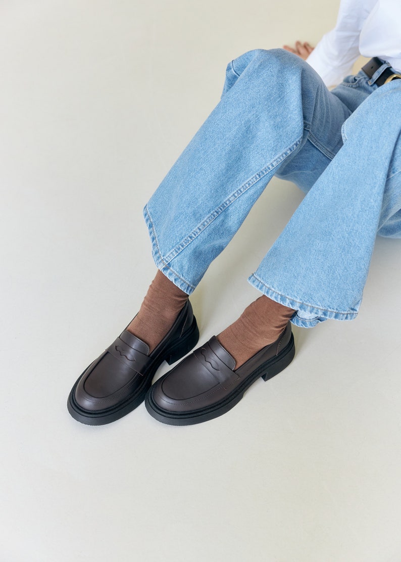 K&ouml;nnte beinhalten: Dunkelbraune Leder-Loafer mit dicker schwarzer Sohle. Die Schuhe haben ein klassisches Penny-Loafer-Design. Die Schuhe werden mit braunen Socken und hellblauen Jeans getragen. Die Schuhe sind etwa 25 cm lang.