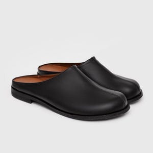 Könnte beinhalten: Ein Paar schwarze Leder-Clogs. Die Schuhe haben eine runde Spitze und eine flache Sohle. Die Innenseite ist hellbraun. Die Clogs werden auf weißem Hintergrund präsentiert.