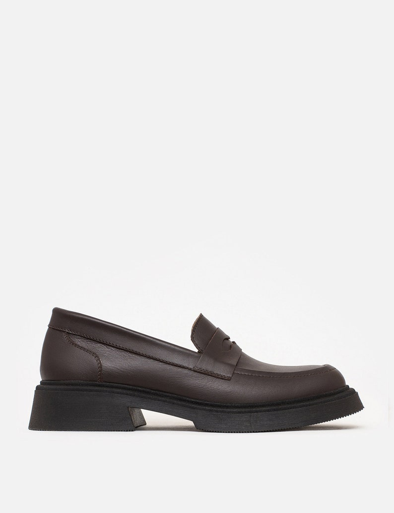 K&ouml;nnte beinhalten: Ein brauner Lederloafer mit einer klobigen schwarzen Sohle. Der Schuh hat ein klassisches Penny-Loafer-Design mit runder Spitze und niedrigem Absatz. Der Schuh ist aus glattem, strukturiertem Leder gefertigt.