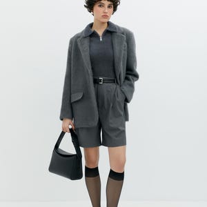 Puede incluir: Una mujer con un blazer gris difuso, una camisa de cuello gris oscuro, pantalones cortos grises, calcetines negros transparentes y zapatos negros. Lleva un bolso negro.
