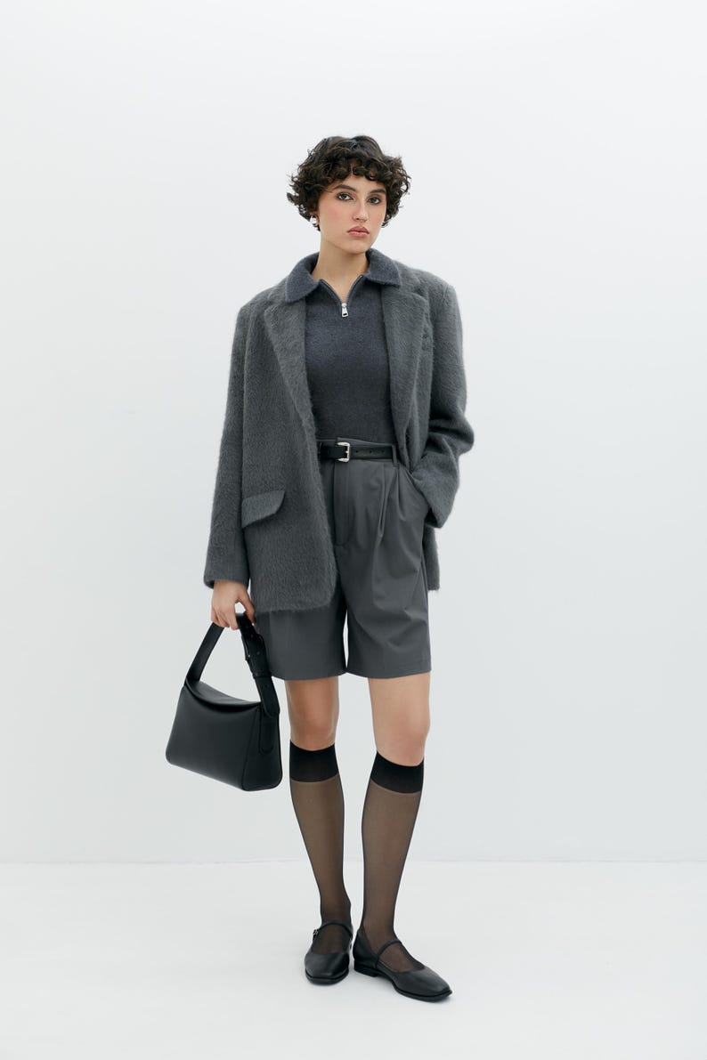 Puede incluir: Una mujer con un blazer gris difuso, una camisa de cuello gris oscuro, pantalones cortos grises, calcetines negros transparentes y zapatos negros. Lleva un bolso negro.
