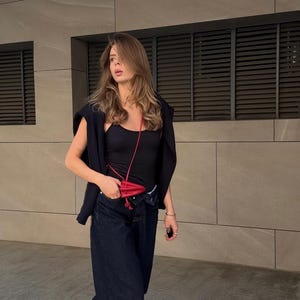 May include: A person wearing a black tank top, navy blue jeans, and a black sweater draped over the shoulders. A small red purse hangs from the neck. Red shoes complete the outfit. The background is a neutral-toned building.