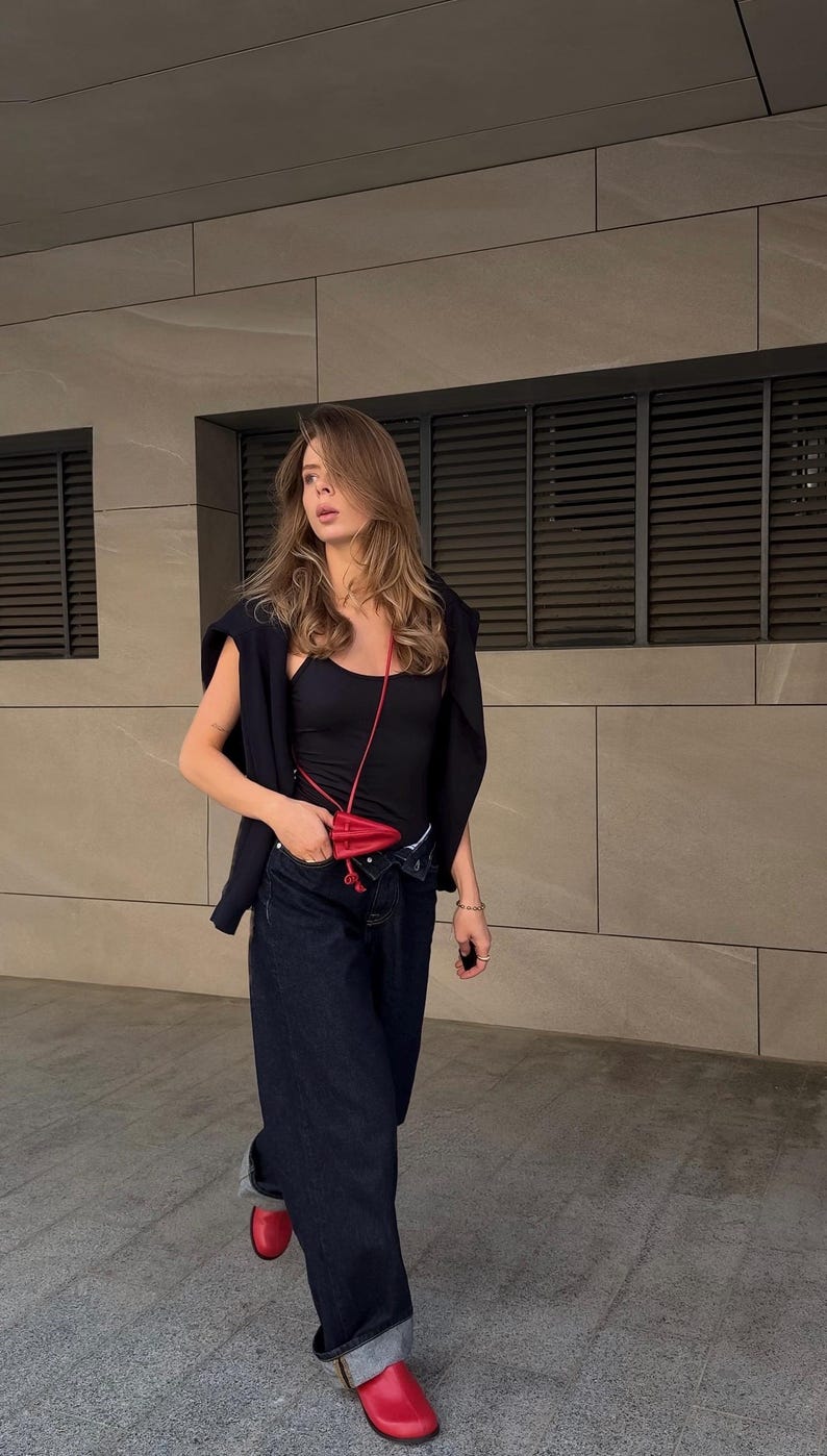 May include: A person wearing a black tank top, navy blue jeans, and a black sweater draped over the shoulders. A small red purse hangs from the neck. Red shoes complete the outfit. The background is a neutral-toned building.