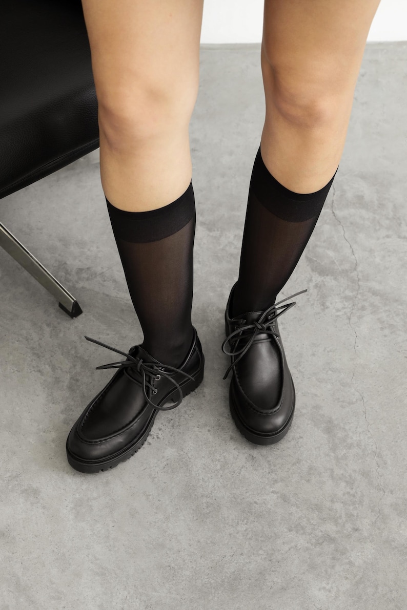 May include: A pair of black leather lace-up shoes with a thick lug sole and a raised seam detail on the toe, worn with sheer black knee-high socks that feature a solid opaque band at the top, providing a clean and uniform appearance.