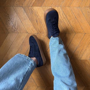 Puede incluir: Zapatillas de color azul marino con suelas marrones. Los zapatos est&aacute;n atados con cordones y se usan con vaqueros de mezclilla azul claro. Los zapatos est&aacute;n sobre un suelo de madera.
