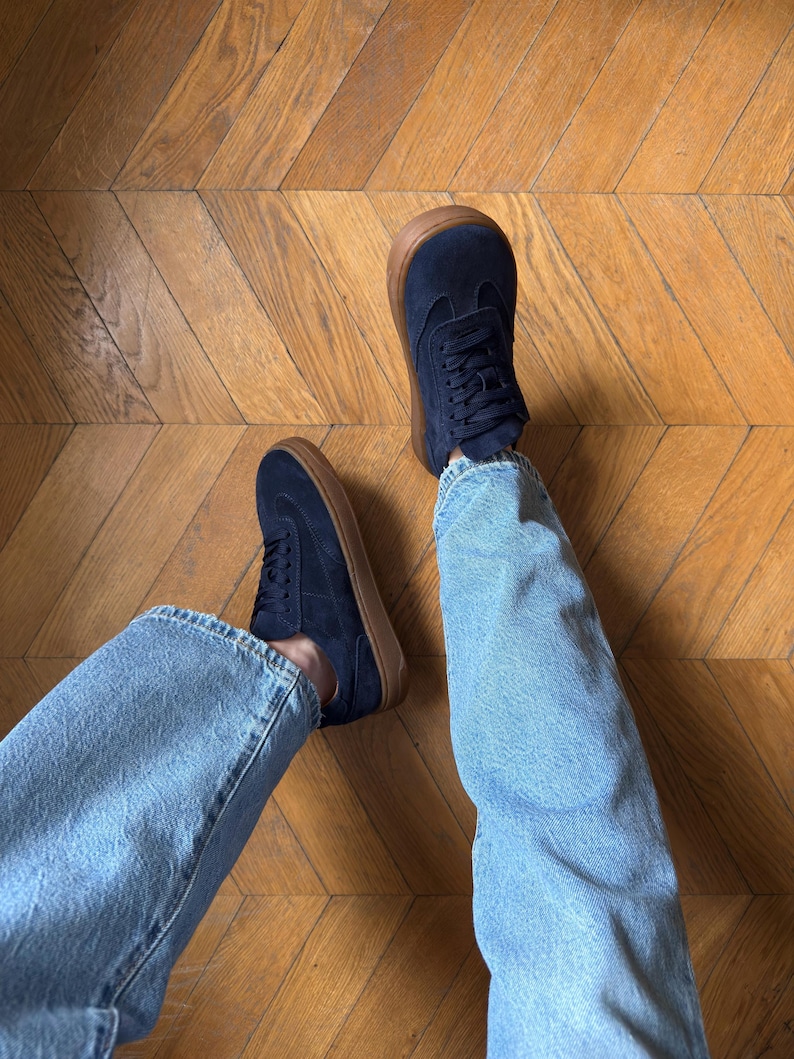 Puede incluir: Zapatillas de color azul marino con suelas marrones. Los zapatos est&aacute;n atados con cordones y se usan con vaqueros de mezclilla azul claro. Los zapatos est&aacute;n sobre un suelo de madera.