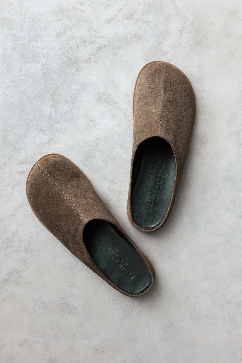 May include: A pair of brown suede slip-on mules with a rounded toe and a centre seam. The interior footbed is dark green with the embossed text LEONCHENKO MADE IN VIETNAM. The soles are a light tan colour, providing a contrast to the brown upper.