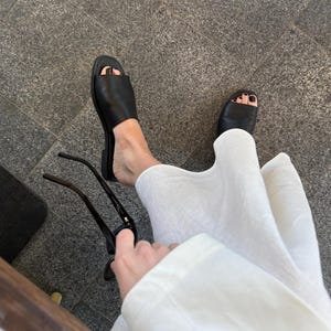 May include: Black leather slide sandals with a square toe. The sandals are worn with a white linen outfit. A pair of black sunglasses are held in the hand.