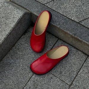 May include: A pair of red leather slip-on shoes with a rounded toe and a black sole. The shoes are displayed on a gray stone surface. The interior of the shoe is a light tan color.