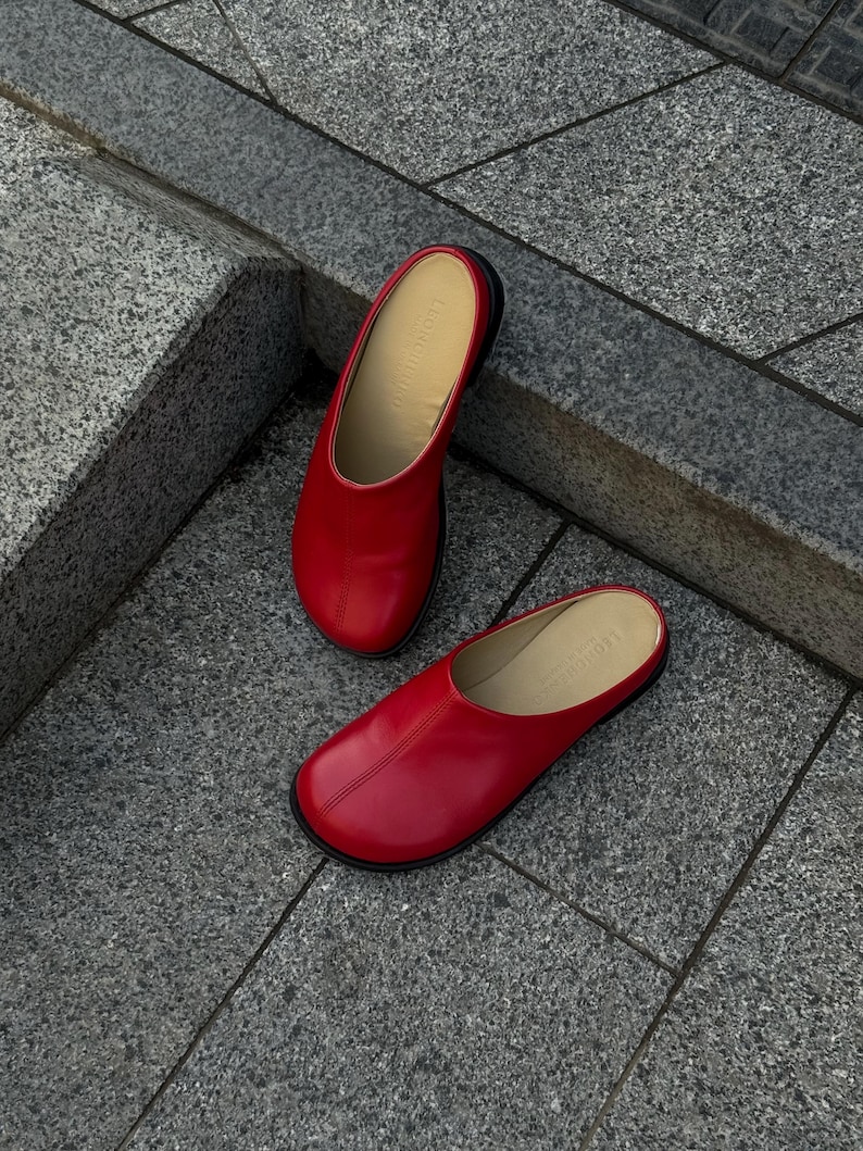 May include: A pair of red leather slip-on shoes with a rounded toe and a black sole. The shoes are displayed on a gray stone surface. The interior of the shoe is a light tan color.