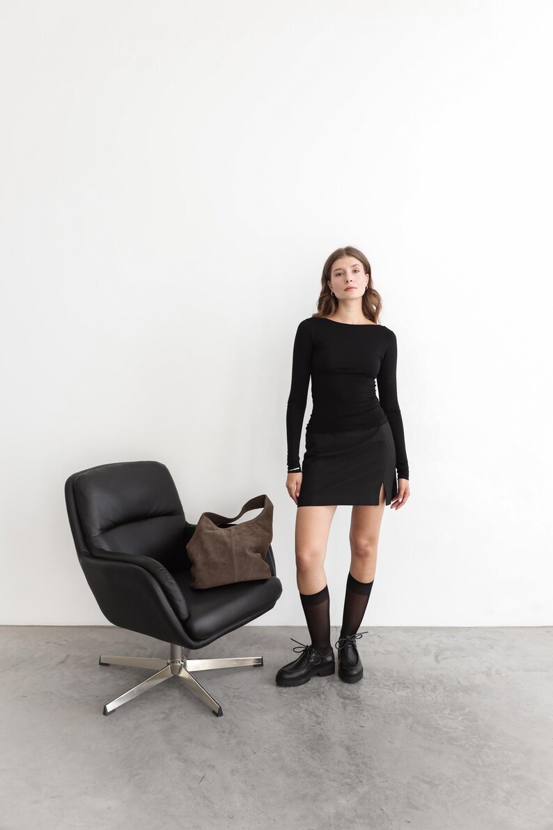 May include: A long-sleeved black top, a black mini skirt with a side slit, sheer black knee-high socks, and black lace-up shoes. A brown suede hobo bag rests on a black leather swivel chair with a silver metal base against a plain white wall.
