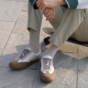 May include: A pair of white and brown sneakers with a low profile. The shoes have a brown suede toe cap and heel, with white leather panels. The soles are brown. The shoes are on a gray stone surface.