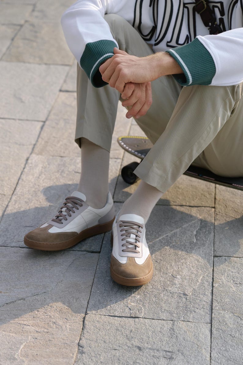 May include: A pair of white and brown sneakers with a low profile. The shoes have a brown suede toe cap and heel, with white leather panels. The soles are brown. The shoes are on a gray stone surface.