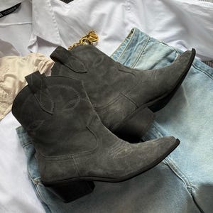 Op de afbeelding: Een paar donkergrijze suède enkellaarsjes met een puntige neus, een gestapelde blokhak en decoratieve ton-sur-ton stiksels op de schacht. De laarsjes liggen op een wit overhemd met kraag en een lichtblauwe spijkerbroek met een gouden ketting.