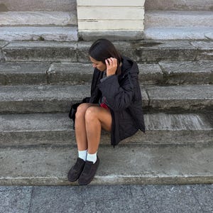 Puede incluir: Una persona con una chaqueta negra acolchada, una falda roja, calcetines blancos y zapatillas deportivas marrones oscuras est&aacute; sentada en escalones de piedra. La chaqueta tiene capucha y un patr&oacute;n de diamantes. Los escalones son de piedra gris.