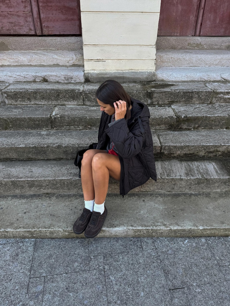 Puede incluir: Una persona con una chaqueta negra acolchada, una falda roja, calcetines blancos y zapatillas deportivas marrones oscuras est&aacute; sentada en escalones de piedra. La chaqueta tiene capucha y un patr&oacute;n de diamantes. Los escalones son de piedra gris.