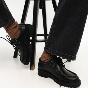 May include: A pair of black leather shoes with a moccasin-style stitched toe, black laces, and a thick, lugged rubber sole. The shoes are worn with brown ribbed socks and dark gray denim jeans, positioned near a black wooden stool.