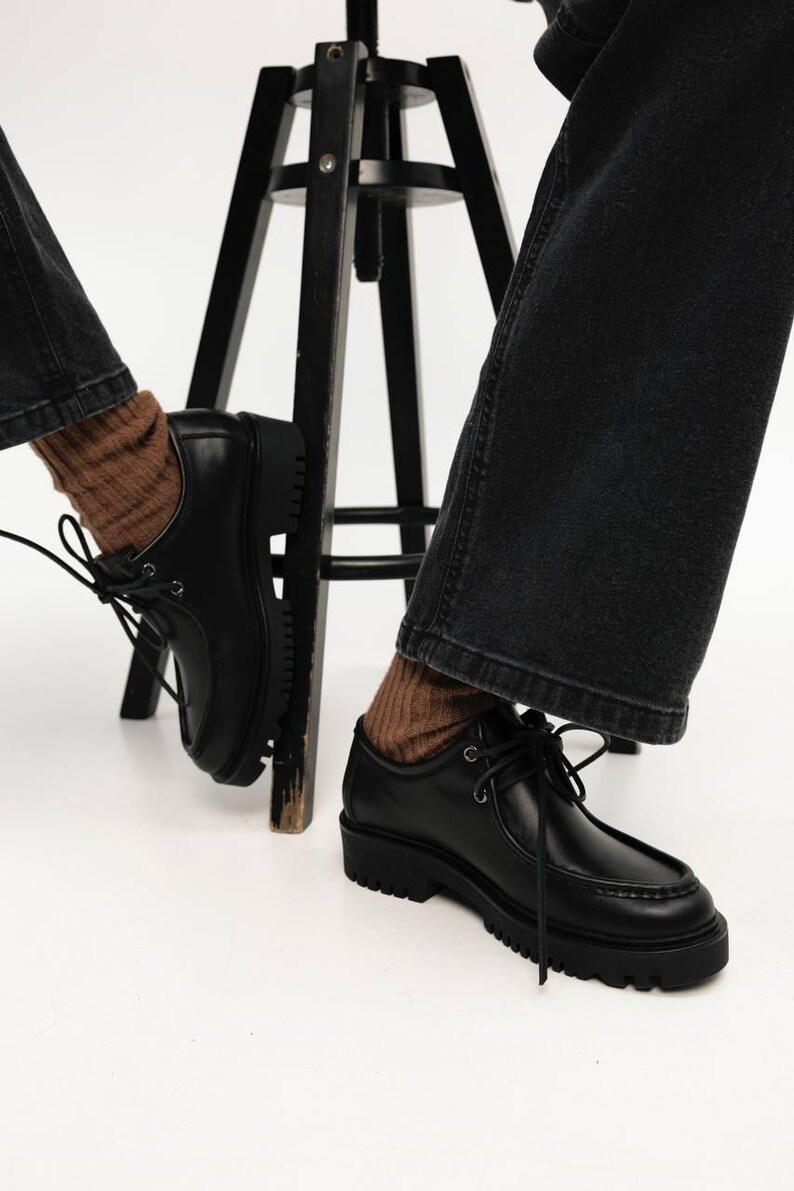 May include: A pair of black leather shoes with a moccasin-style stitched toe, black laces, and a thick, lugged rubber sole. The shoes are worn with brown ribbed socks and dark gray denim jeans, positioned near a black wooden stool.