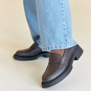 K&ouml;nnte beinhalten: Braune Leder-Loafer mit einer klobigen schwarzen Sohle. Die Loafer haben ein klassisches Penny-Loafer-Design mit einem dekorativen Detail auf dem Blatt. Die Schuhe werden mit hellblauen Jeans kombiniert.