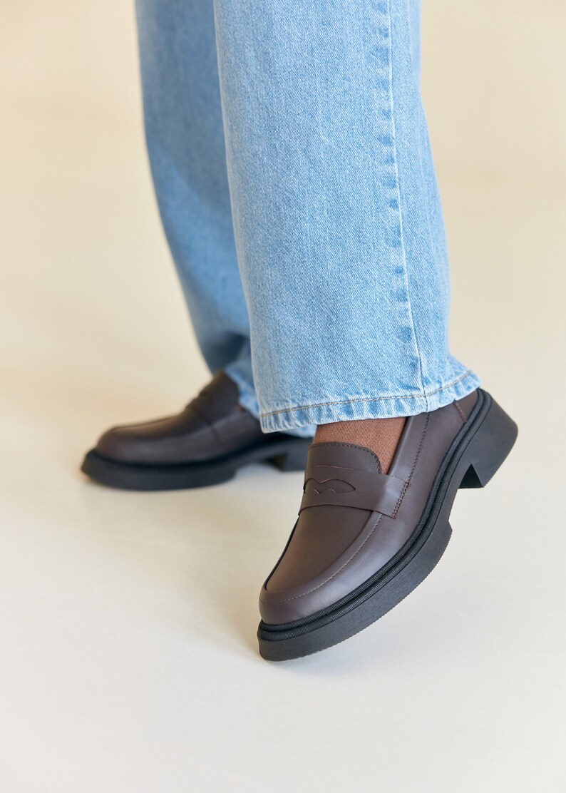 K&ouml;nnte beinhalten: Braune Leder-Loafer mit einer klobigen schwarzen Sohle. Die Loafer haben ein klassisches Penny-Loafer-Design mit einem dekorativen Detail auf dem Blatt. Die Schuhe werden mit hellblauen Jeans kombiniert.