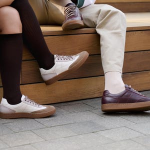 May include: A collection of sneakers in various styles. One pair features a white and tan color scheme with a brown sole. Another pair is a deep burgundy with a brown sole. A third pair is visible in the background.