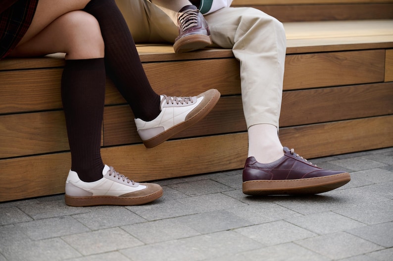 May include: A collection of sneakers in various styles. One pair features a white and tan color scheme with a brown sole. Another pair is a deep burgundy with a brown sole. A third pair is visible in the background.