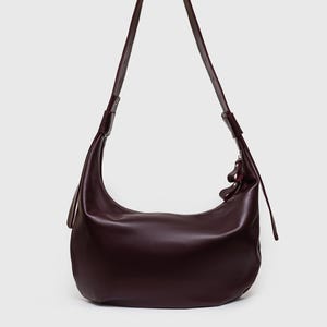 May include: A deep burgundy shoulder bag with a curved, crescent shape. The bag features a long, thin strap and a zipper closure. The leather bag has a smooth, polished finish and a minimalist design.