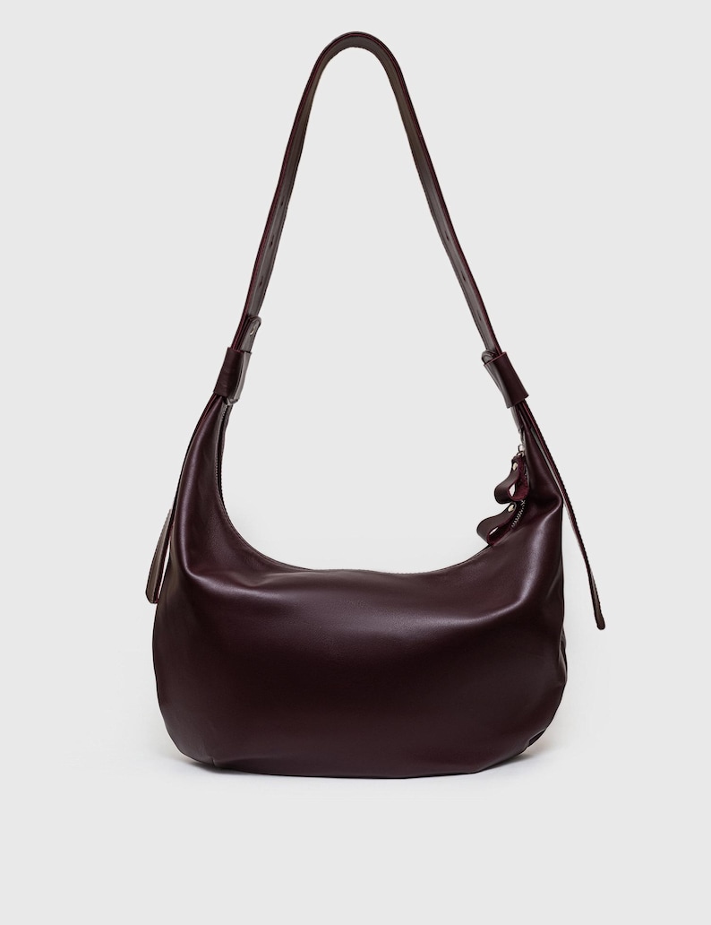 May include: A deep burgundy shoulder bag with a curved, crescent shape. The bag features a long, thin strap and a zipper closure. The leather bag has a smooth, polished finish and a minimalist design.