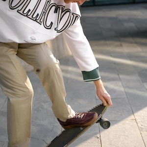 May include: A person wearing a white sweatshirt with "COLLECTOR" printed in black and tan, green trim, and tan pants, stepping onto a skateboard. The shoes are burgundy.
