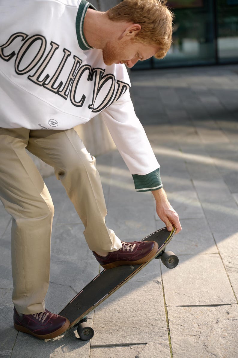 May include: A person wearing a white sweatshirt with "COLLECTOR" printed in black and tan, green trim, and tan pants, stepping onto a skateboard. The shoes are burgundy.