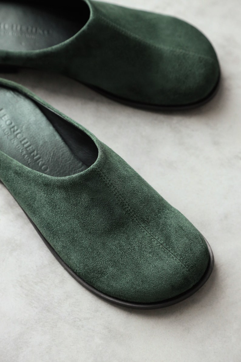 May include: A pair of dark green suede slip-on shoes with a rounded toe and a centre seam. The interior footbed is black and features the embossed text "LEONCHENKO" and "MADE IN UKRAINE" near the heel area, with a thin black outer sole.