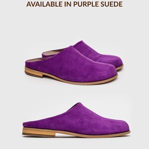 May include: A pair of purple suede mules with a rounded toe and a low, flat wooden heel. The text AVAILABLE IN PURPLE SUEDE is printed above the shoes, and the text *also available in other colours is printed below the shoes.