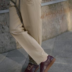 May include: Khaki-colored pants paired with burgundy leather sneakers with brown soles. The sneakers have a low-profile design and lace-up closure. The pants are straight-legged and have a classic, casual style. The outfit is suitable for everyday wear.