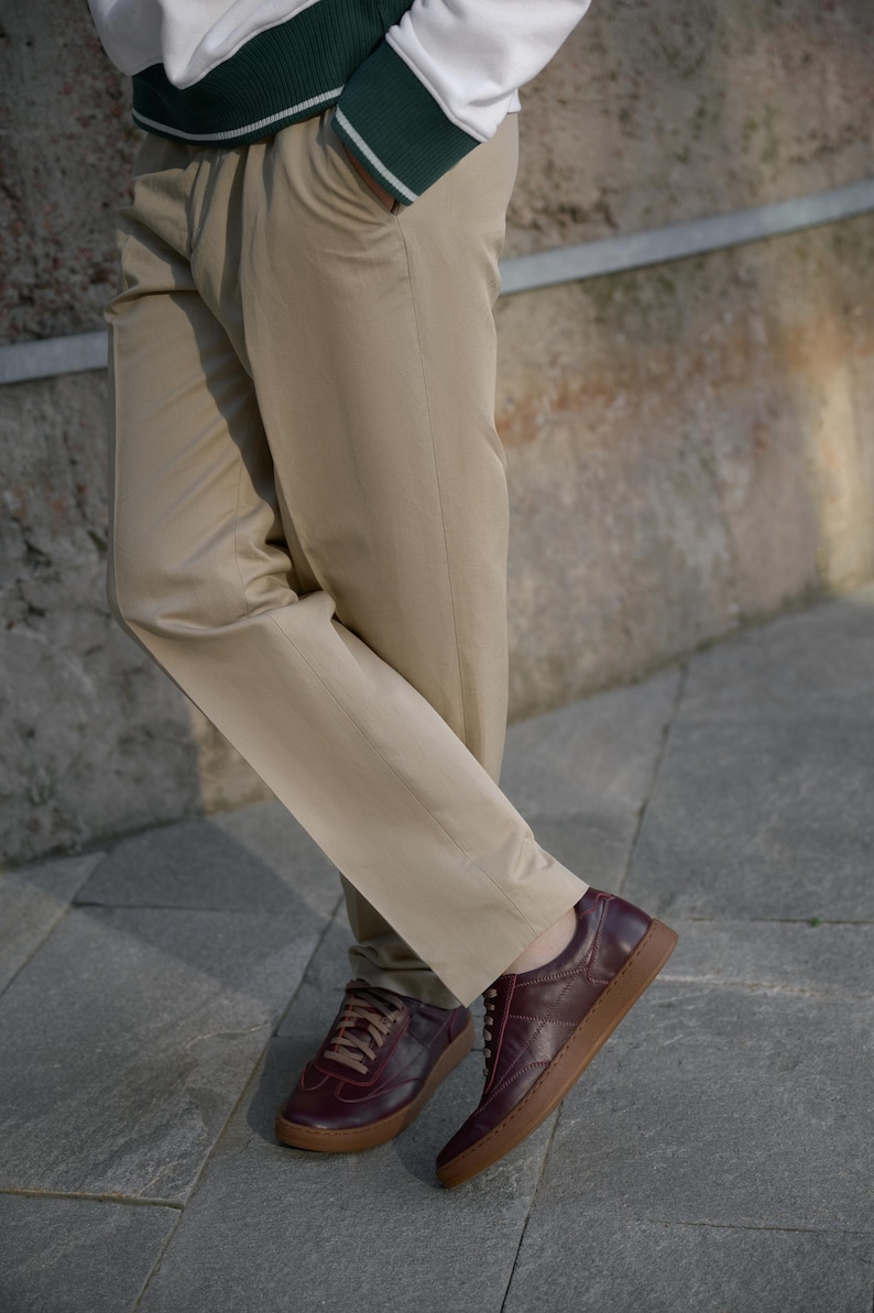 May include: Khaki-colored pants paired with burgundy leather sneakers with brown soles. The sneakers have a low-profile design and lace-up closure. The pants are straight-legged and have a classic, casual style. The outfit is suitable for everyday wear.