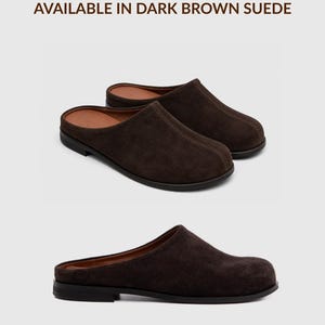 May include: A pair of dark brown suede mules with a rounded toe, a low heel, and a tan interior lining. The text at the top reads AVAILABLE IN DARK BROWN SUEDE and the text at the bottom reads *also available in other colours.