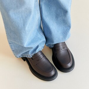 K&ouml;nnte beinhalten: Ein Paar dunkelbraune Leder-Loafer mit dicker schwarzer Sohle. Die Schuhe haben ein klassisches Penny-Loafer-Design und werden mit hellblauen Jeans getragen.