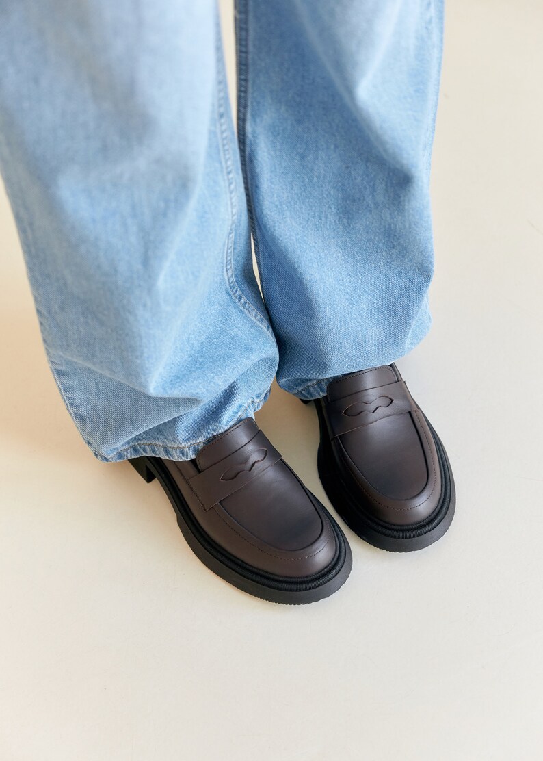 K&ouml;nnte beinhalten: Ein Paar dunkelbraune Leder-Loafer mit dicker schwarzer Sohle. Die Schuhe haben ein klassisches Penny-Loafer-Design und werden mit hellblauen Jeans getragen.