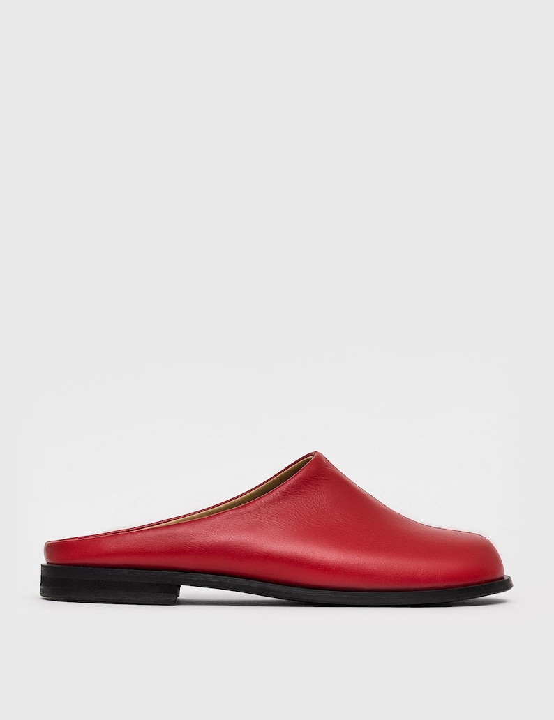 May include: A single red leather mule with a rounded toe and a low, black sole. The shoe has a simple, minimalist design and a smooth texture. The heel is approximately 1 inch high.