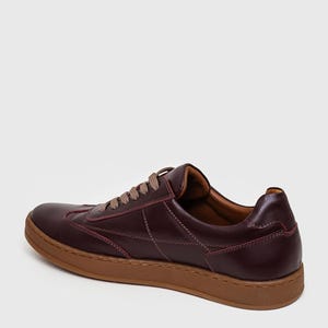 May include: Burgundy leather sneakers with brown soles and laces. The shoes feature stitched detailing and a classic low-top design. The sneakers are a stylish choice for casual wear.