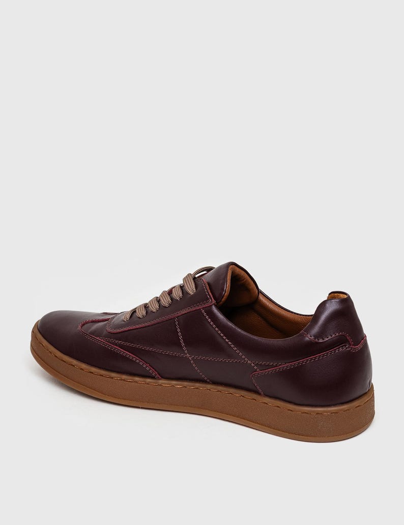 May include: Burgundy leather sneakers with brown soles and laces. The shoes feature stitched detailing and a classic low-top design. The sneakers are a stylish choice for casual wear.