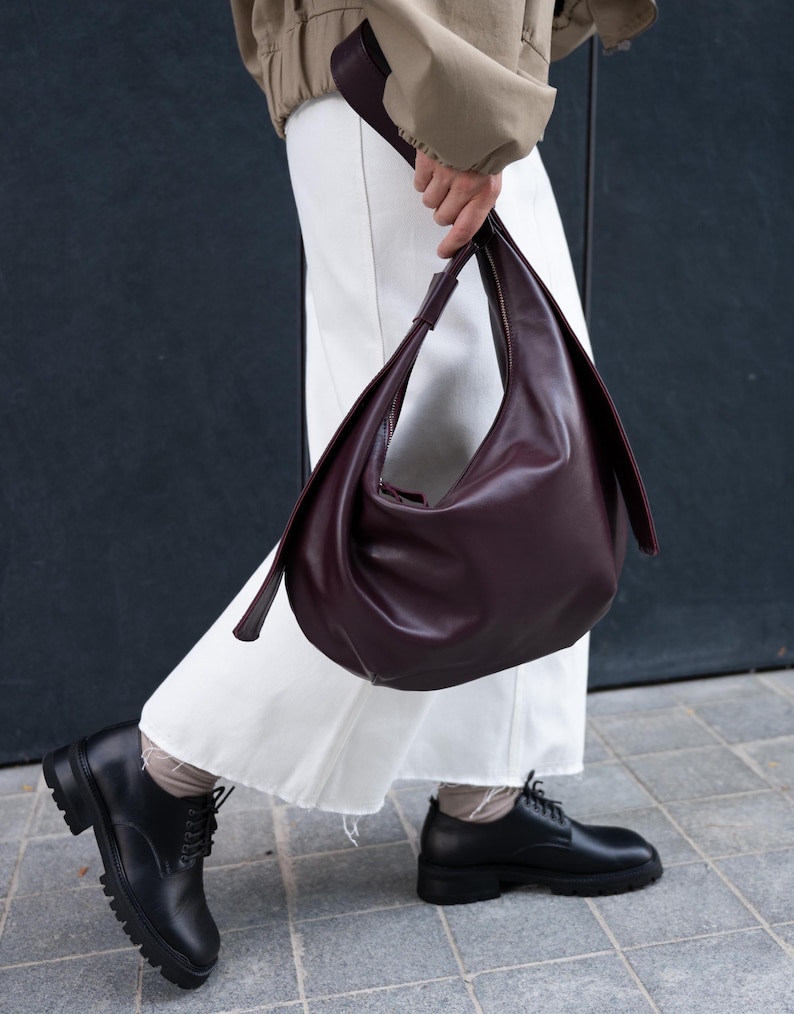 May include: A burgundy leather shoulder bag with a curved design. The bag has a long strap and a zipper closure. The person is wearing white pants and black shoes with thick soles. The background is a dark wall.