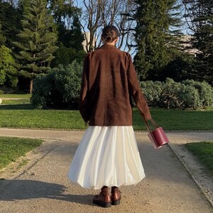May include: A brown blazer worn over a voluminous white midi skirt, paired with dark brown ankle boots and a cylindrical burgundy handbag. The outfit is captured from behind, showcasing the contrast between the structured jacket and the skirt.