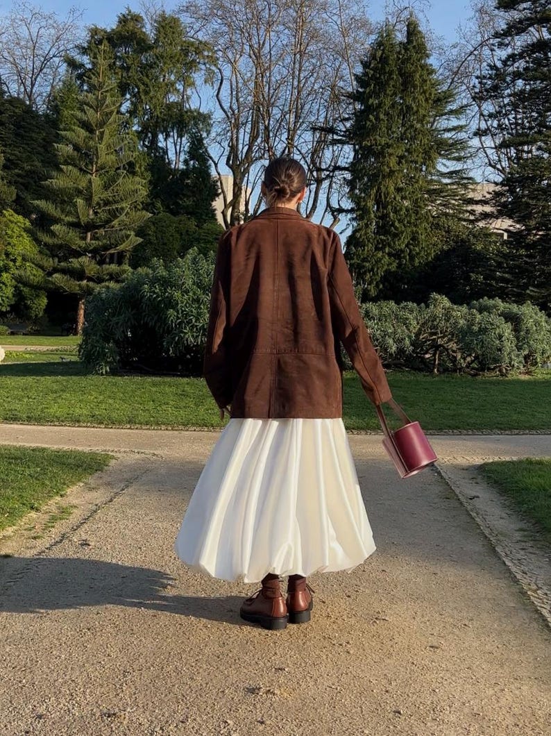 May include: A brown blazer worn over a voluminous white midi skirt, paired with dark brown ankle boots and a cylindrical burgundy handbag. The outfit is captured from behind, showcasing the contrast between the structured jacket and the skirt.
