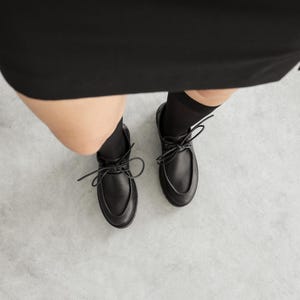 May include: A pair of black leather shoes with a rounded toe, a raised seam along the top, and black laces tied in a bow. The shoes are worn with black socks and are positioned below a black garment, showing a clear view of the footwear design.