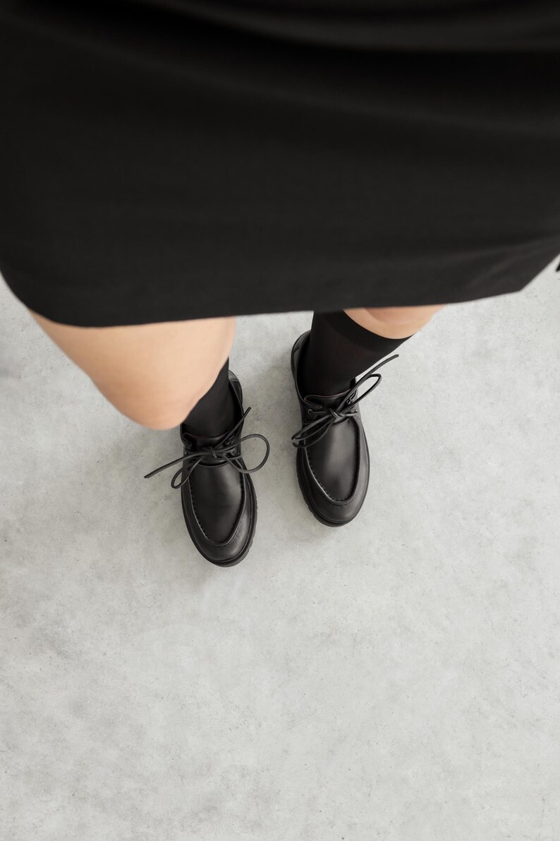 May include: A pair of black leather shoes with a rounded toe, a raised seam along the top, and black laces tied in a bow. The shoes are worn with black socks and are positioned below a black garment, showing a clear view of the footwear design.