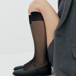 Puede incluir: Zapatillas negras de cuero con una sola correa y hebilla. Los zapatos se llevan con medias negras transparentes.
