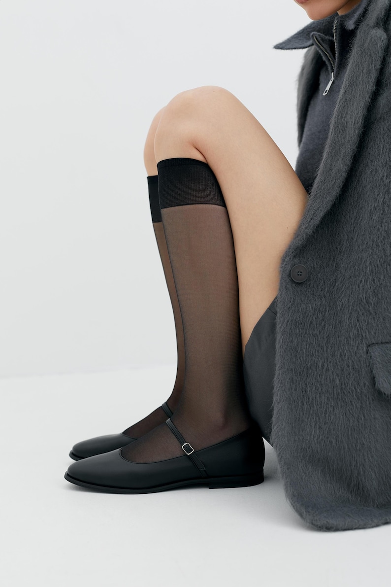 Puede incluir: Zapatillas negras de cuero con una sola correa y hebilla. Los zapatos se llevan con medias negras transparentes.