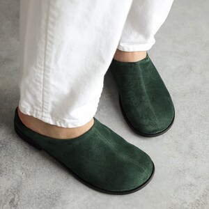 May include: A pair of dark forest green suede slip-on mules featuring a rounded toe, a central seam detail running down the vamp, and a thin black sole. The footwear has an open-back design and a soft, matte texture across the upper surface.