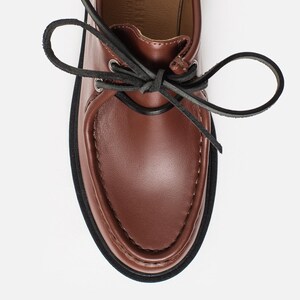 May include: A brown leather shoe with a moccasin-style toe, featuring black leather laces tied in a bow through silver-toned metal eyelets. The shoe has a thick black sole and a tan interior lining with the text ONCHENI MADE IN UKRAINE visible.